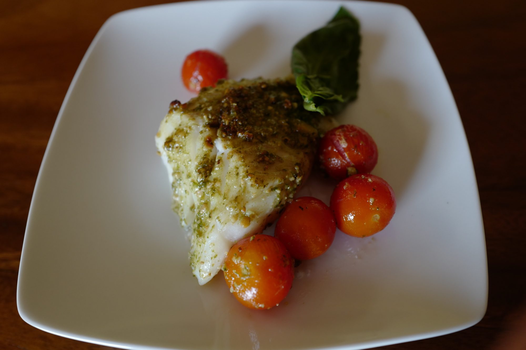 pesto baked cod with cherry tomatoes, basil leaf, on a white plate by foodjoya